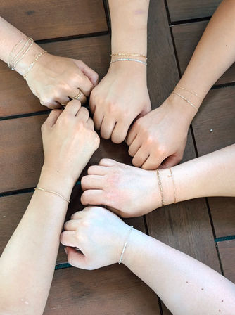 Six hands in a circle on a wooden table, each wrist wearing delicate gold and silver chain bracelets and a few minimalist rings — group bracelet styling shot