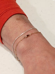 Close-up of a wrist in an orange sleeve wearing stacked delicate silver and thin gold chain bracelets with a small clasp on a white speckled countertop