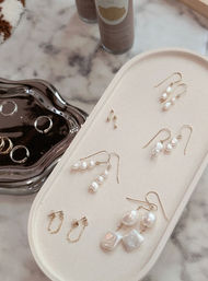 Stylish flat-lay of delicate pearl drop and gold hoop earrings on a cream oval jewelry tray beside a mirrored ring dish and skincare bottles on a marble countertop