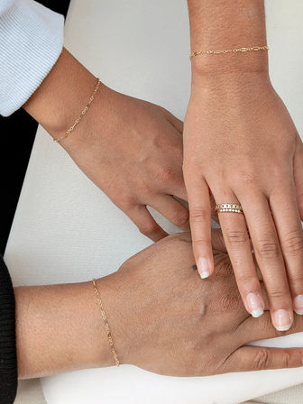 Close-up of three hands layered on a white surface wearing delicate gold chain bracelets and a stacked band ring, showcasing minimalist jewelry and light manicures.