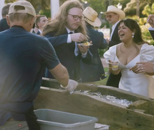 Bride in white gown laughing with guests at an outdoor wedding seafood station — man in suit with long hair eats oysters from an ice-filled wooden bar while others hold drinks.