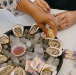 Close-up of fresh shucked oysters on ice in a black platter with a hand squeezing lemon, a cup of cocktail sauce and wrapped crackers on a white table.