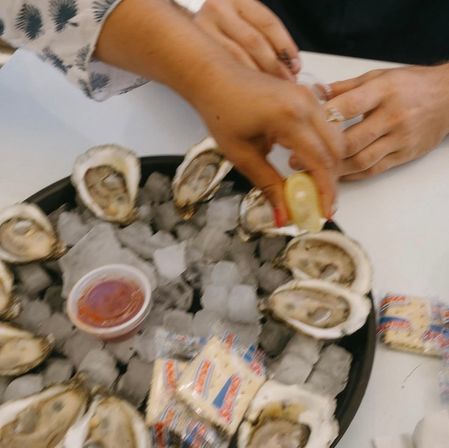 Close-up of fresh shucked oysters on ice in a black platter with a hand squeezing lemon, a cup of cocktail sauce and wrapped crackers on a white table.