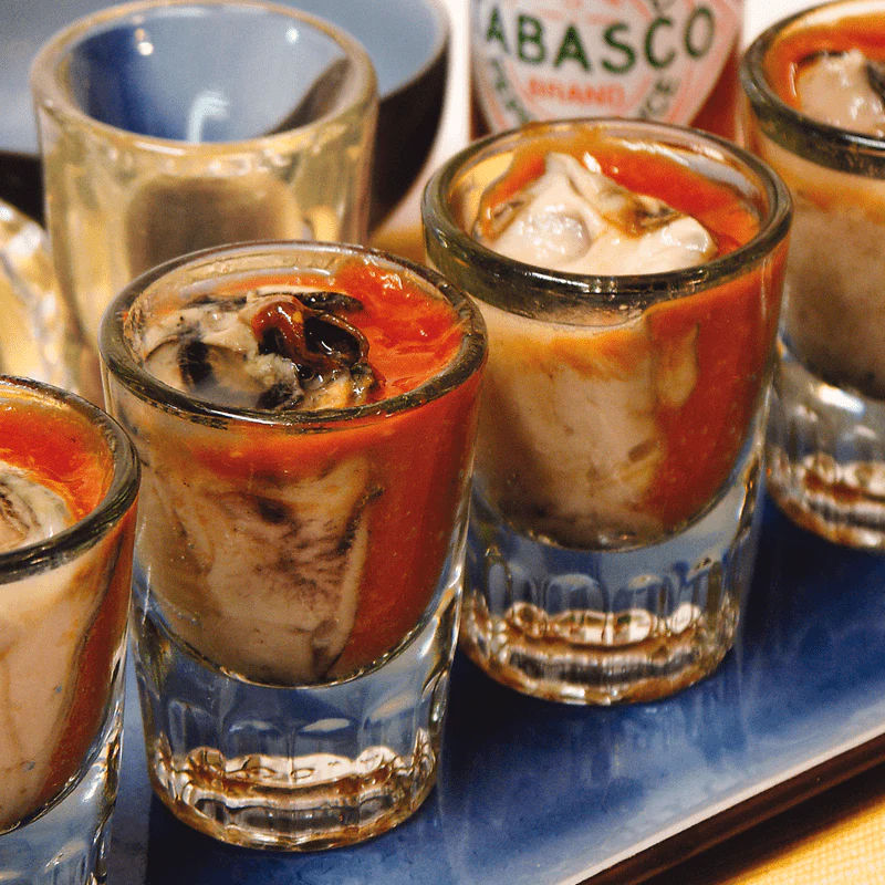 Oyster shooters — raw oysters in shot glasses splashed with red cocktail and hot sauce on a blue serving tray