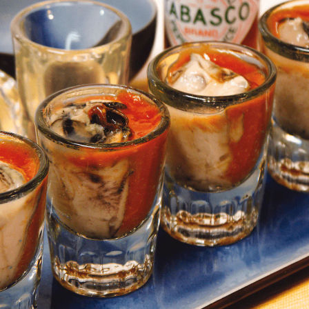 Oyster shooters — raw oysters in shot glasses splashed with red cocktail and hot sauce on a blue serving tray