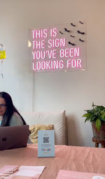 Playful pink neon sign reading "THIS IS THE SIGN YOU'VE BEEN LOOKING FOR" above a cozy wellness studio reception with a laptop, QR review card, potted plant and pink decor.