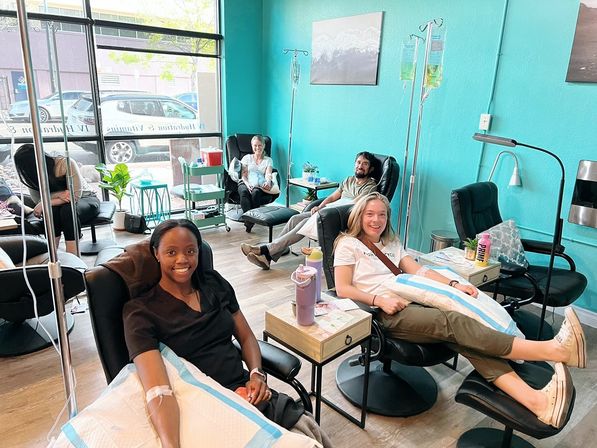 Smiling adults relaxing in reclining chairs at a bright teal IV therapy wellness lounge, receiving hydration infusions from IV poles in a cozy, modern clinic setting.