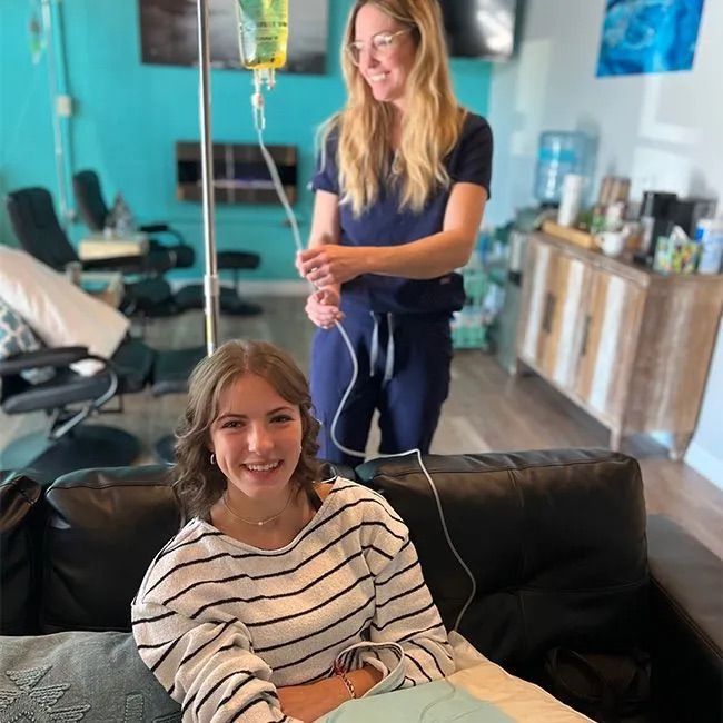 Smiling young woman relaxed on a black leather sofa receiving a hydration IV drip as a clinician adjusts the IV pole in a bright teal wellness clinic lounge with recliners and a refreshment area.