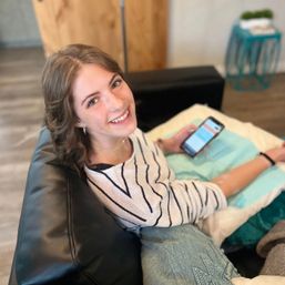 Smiling woman in a striped sweater lounging on a black leather couch in a cozy living room, relaxing with a smartphone on a lap pillow.