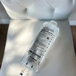 Clear 1,000 mL 0.9% sodium chloride IV saline bag resting on a white tufted chair, medical infusion bag with printed label and visible drip ports in soft light