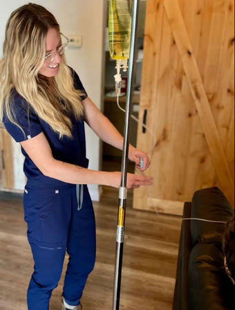 Smiling healthcare professional in navy scrubs adjusting an IV pole during a home infusion visit next to a leather couch and wooden barn door
