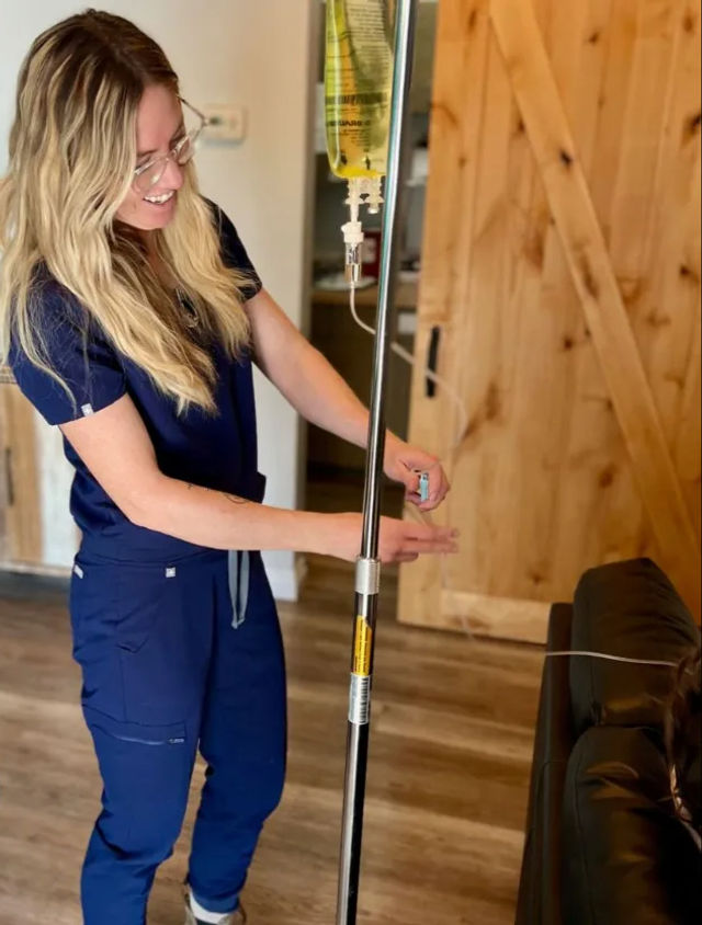 Smiling healthcare professional in navy scrubs adjusting an IV pole during a home infusion visit next to a leather couch and wooden barn door