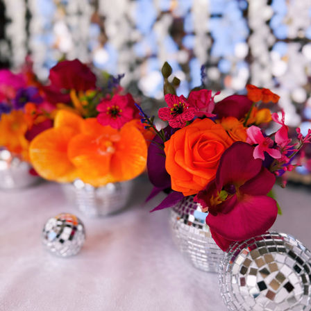 Vibrant event centerpiece with bright orange rose, magenta orchids and mirrored disco-ball vases on a white tablecloth and sparkly sequin backdrop