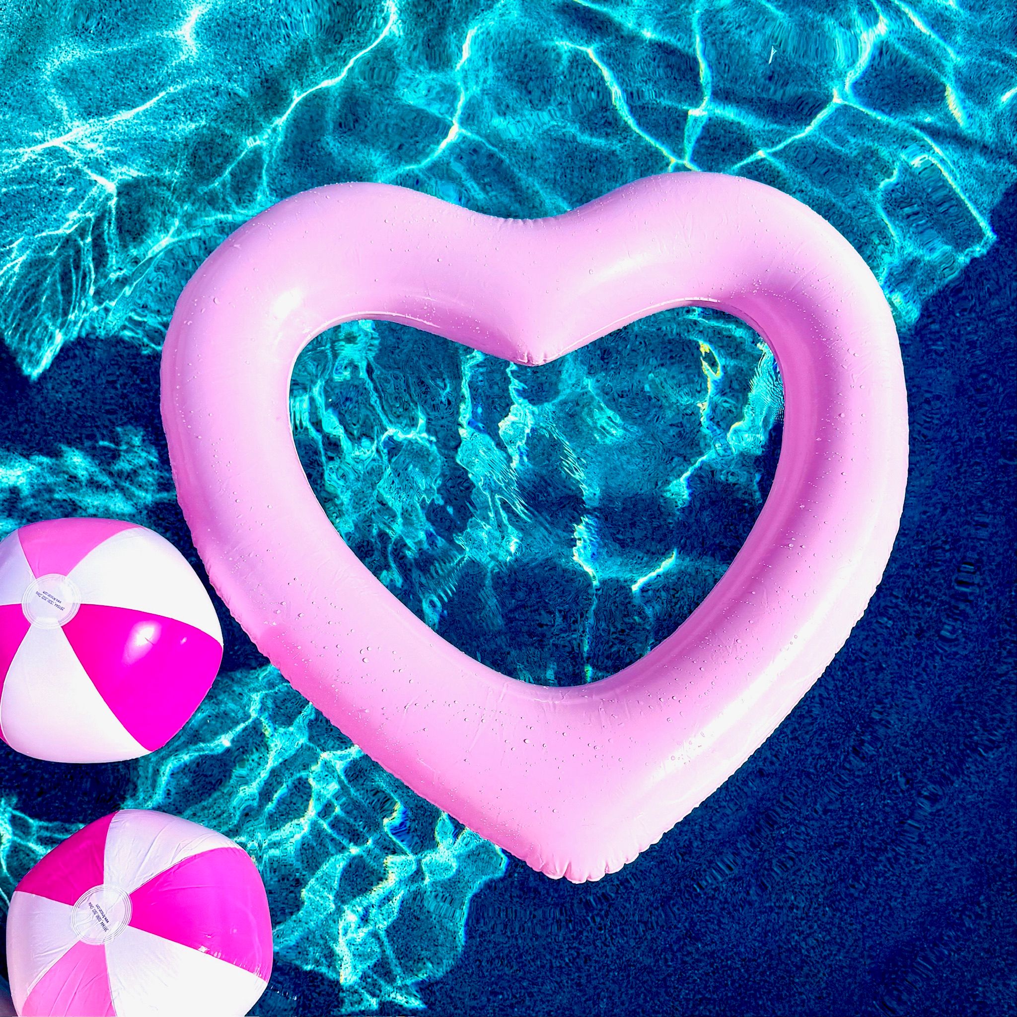 Pink heart-shaped inflatable and two pink-and-white beach balls floating on turquoise swimming pool water in a sunlit summer pool scene
