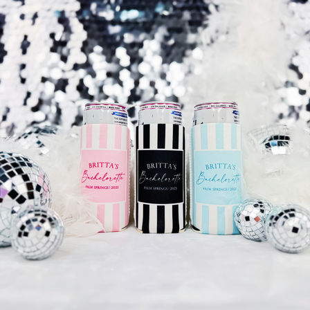 Three striped can sleeves in pink, black, and blue labeled bachelorette (Palm Springs 2025) arranged with white feathers, disco balls and a sparkling sequin backdrop, party-ready