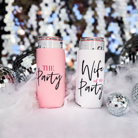 Two slim can coolers on a feather-covered party table, pink labeled The Party and white labeled Wife of the Party, surrounded by mirrored disco balls and a sparkling bokeh backdrop — playful bachelorette or bridal shower decor.