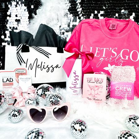 Sparkling pink bachelorette gift set with personalized tote and tumbler, bright pink Lets Go Girls tee, cozy crew socks, heart-shaped sunglasses and mini disco balls on a sequin backdrop