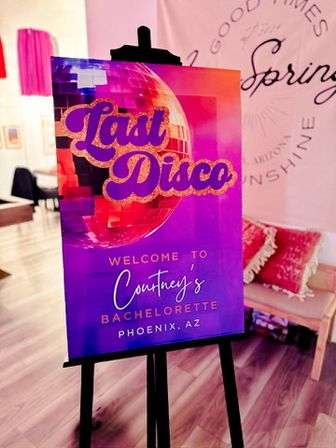 Vibrant purple-pink easel sign with a glittery disco ball and bold “Last Disco” text, welcoming guests to a bachelorette party in Phoenix, AZ in a cozy studio setting.