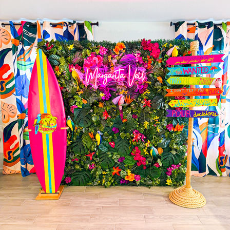 Bright tropical photo booth backdrop: lush faux green wall with colorful orchids and parrots, pink neon margarita sign, striped pink surfboard on a wooden stand, and rope-mounted beach signpost with playful directional arrows — indoor beach-themed party decor.