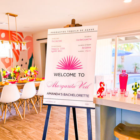 Margarita-themed bachelorette party setup in Scottsdale, AZ with an easel welcome sign, long dining table dressed with bright floral centerpieces, a pink cocktail station with shot glasses and playful flamingo and cactus accents, and a pool visible through sliding glass doors.
