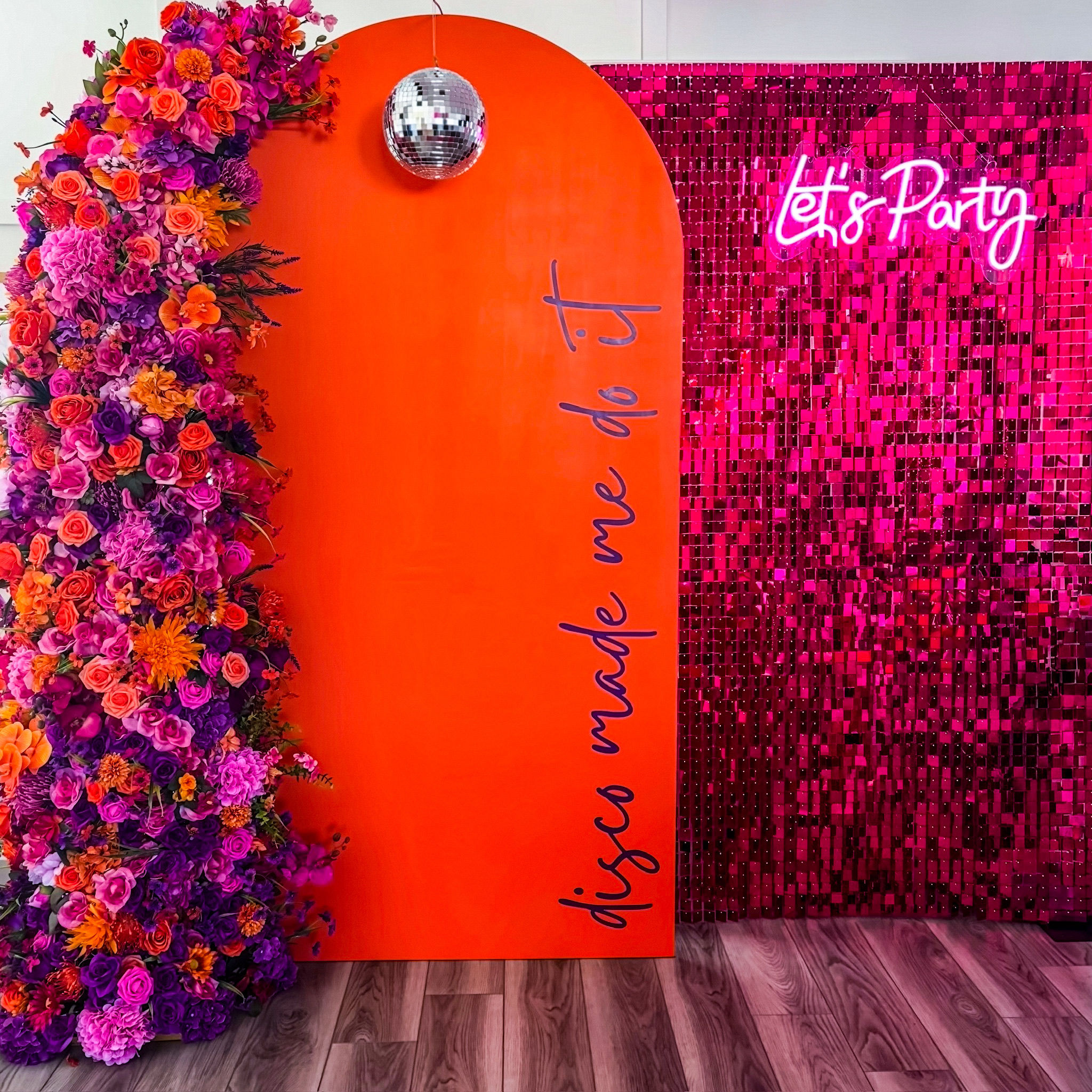 Vibrant party photo backdrop with an orange arched panel reading “disco made me do it,” a hanging silver disco ball, a lush pink‑purple‑orange floral arch on the left, and a hot‑pink sequin wall with neon “Let’s Party” sign over a wood floor.