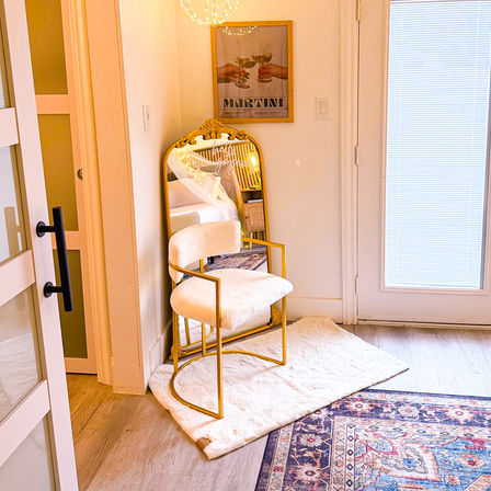 Sunlit cozy bedroom corner with a gold-framed plush chair on a white faux-fur rug, ornate leaning mirror, martini poster and patterned area rug.