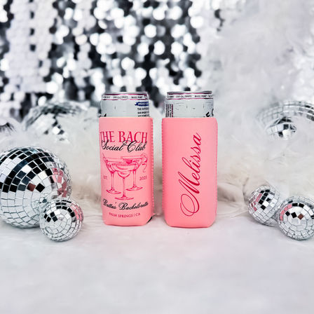 Two pink bachelorette can coolers on fluffy white fur, surrounded by mirrored disco balls and a sparkling sequin backdrop for a Palm Springs party vibe.