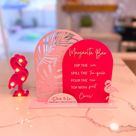 Bright pink tabletop margarita bar sign with tropical leaf cutout, flamingo marquee light, and cocktail instructions for a bachelorette party on a marble countertop.