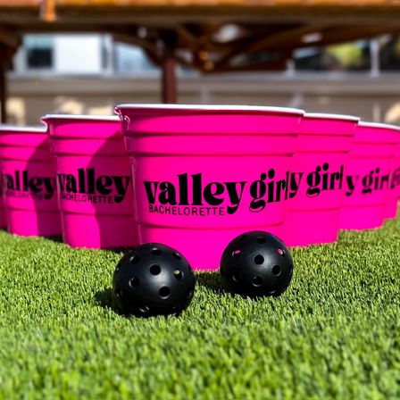 Bright pink plastic party buckets labeled 'bachelorette' lined up on green artificial turf with two black perforated balls — playful outdoor bachelorette party game setup.