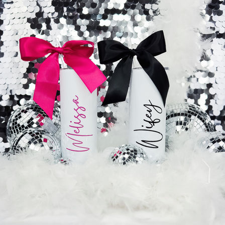 Two tall white personalized tumblers with hot pink and black bows among mirrored disco balls, white feather boa and silver sequin backdrop — glam party favor display.