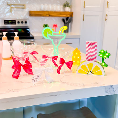 Bright kitchen island party setup with empty margarita glasses tied with pink and white bows, neon cocktail and fruit lights, striped straws and a lemon-themed napkin holder on a white marble countertop