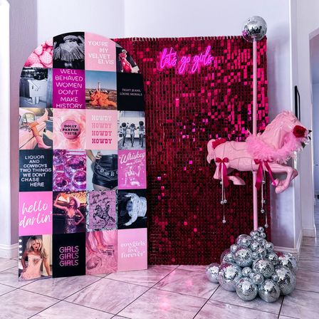Indoor pink party photo booth with shimmering sequin backdrop, neon "let's go girls" sign, pink carousel horse with feather boa, stacked disco balls, and a pink collage panel of retro women-themed graphics.