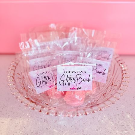 Sparkly pink cotton candy 'Glitter Bomb' party favors, individually wrapped and arranged in a decorative clear glass bowl on a white speckled countertop against a soft pink backdrop