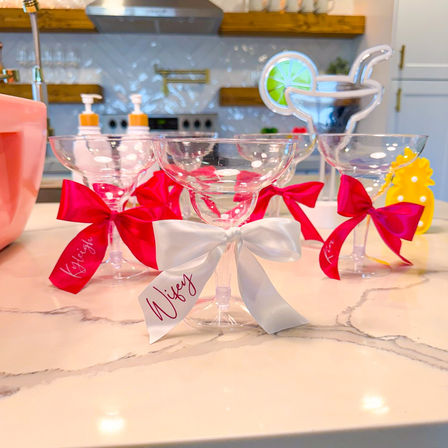 Clear margarita glasses tied with bright pink bows and one white ribbon, arranged on a marble kitchen island as playful cocktail party favors with a blurred stove and lime-shaped decor in the background.