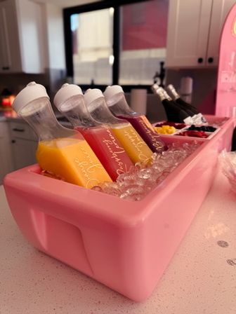 Pink ice-filled cooler on a kitchen counter holding tilted clear bottles of orange, pink, and purple juices with champagne bottles and fruit garnishes — playful brunch mimosa bar setup.