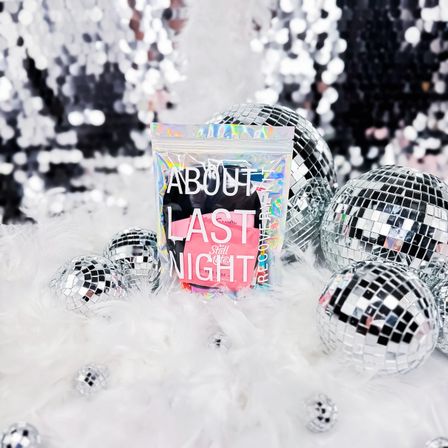 Glam product shot: holographic pouch reading "About Last Night" with pink recovery items on white faux fur, surrounded by mirrored disco balls and a silver sequin backdrop.