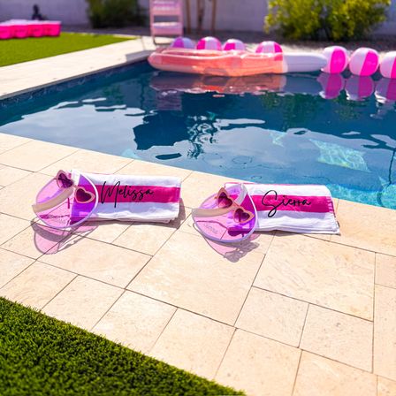Sunny backyard poolside with two pink-and-white beach towels, matching heart-shaped pink sunglasses, and pink inflatable floaties reflected in the blue water — playful summer setup.