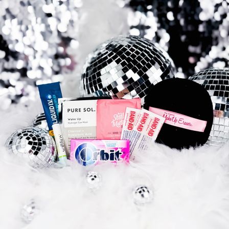 Glam party flatlay of travel-size beauty essentials and first-aid items on white faux fur with reflective disco balls
