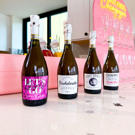 Four mini champagne bottles with playful bachelorette labels lined up on a white counter against a pink bar backdrop with glassware and confetti — festive bachelorette party setup.