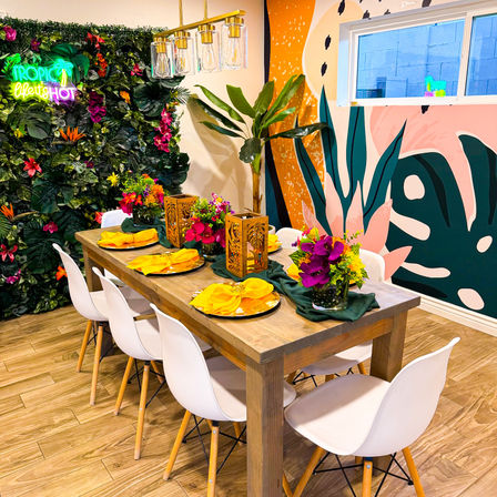 Bright tropical-themed indoor dining area set for six: wooden table with white chairs, yellow napkins, vibrant floral centerpieces, decorative lanterns, leafy green wall with neon accent and colorful mural.