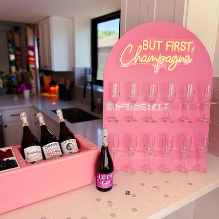 Playful pink champagne station on a bright kitchen counter with neon “But First, Champagne” sign, a pink ice tub holding chilled bottles and berries, an extra bottle on the counter, and rows of clear champagne flutes ready for a party.
