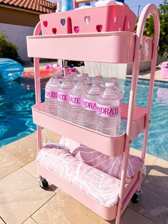 Pink three-tier rolling cart with heart cutouts stocked with bottled water and rolled towels poolside at a sunny backyard