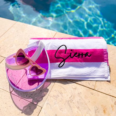 Poolside on a sunlit stone deck: pink-and-white striped personalized towel, translucent pink visor and heart-shaped sunglasses beside a sparkling blue swimming pool.