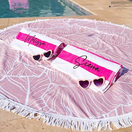 Poolside setup with a round pink leaf-print beach blanket with fringe, two folded pink-and-white striped personalized towels showing handwritten-style names, and matching pink heart-shaped sunglasses.