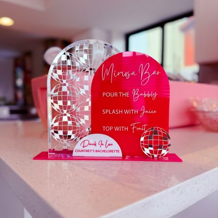 Bright pink acrylic mimosa bar sign with mirrored disco-ball accents and serving instructions, displayed on a white kitchen counter for a bachelorette brunch.