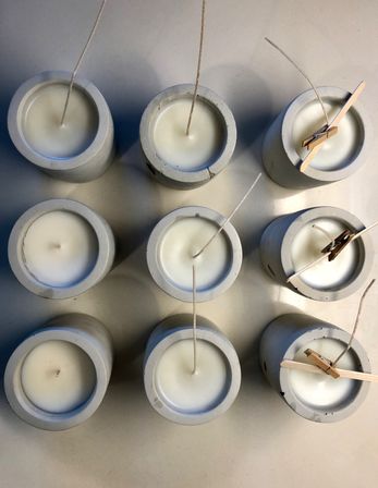 Top-down view of nine poured white candles in round concrete holders arranged in a grid, wicks centered with a few clipped by small clothespins on a neutral surface