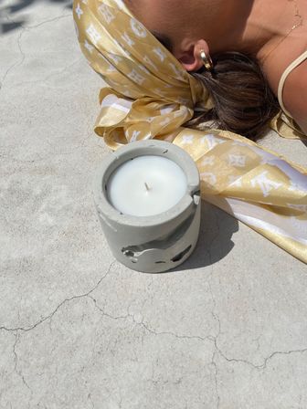 Sunlit outdoor scene with a white candle in a gray concrete holder on cracked concrete beside a reclining person wearing a yellow patterned silk headscarf, gold hoop earring and delicate necklace.