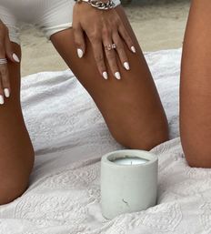 Beach picnic close-up of sun-kissed legs and hands with white manicure, gold rings and chain bracelet resting on a textured white blanket beside a concrete candle on sand