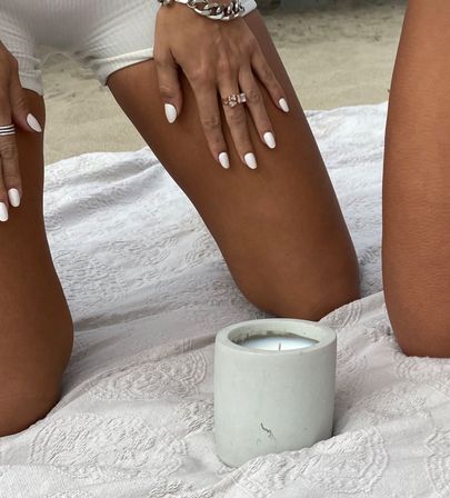Beach picnic close-up of sun-kissed legs and hands with white manicure, gold rings and chain bracelet resting on a textured white blanket beside a concrete candle on sand