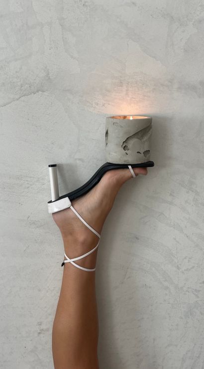 Leg in white strappy heel balancing a lit concrete candle holder against a textured plaster wall, minimalist fashion and home decor composition
